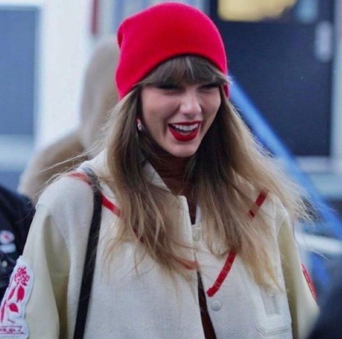 Taylor Swift smiling while wearing a red beanie and cream jacket outdoorsTitle: Taylor Swift in Red Beanie Smiling – May 2025
